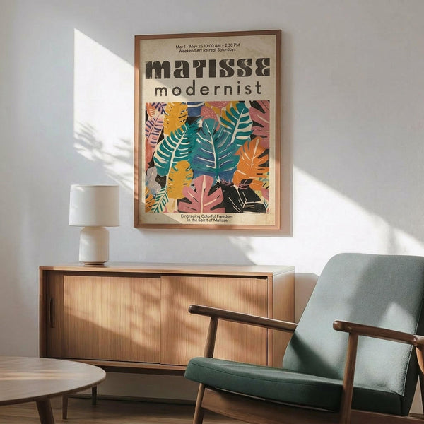 May include: Colorful modernist botanical poster with vibrant cut-out style leaves in teal, orange, pink and yellow, displayed in wooden frame above natural wood sideboard. Mid-century modern room setting features sage green upholstered chair with wooden arms, white ceramic table lamp, and warm sunlight casting geometric shadows on white wall.