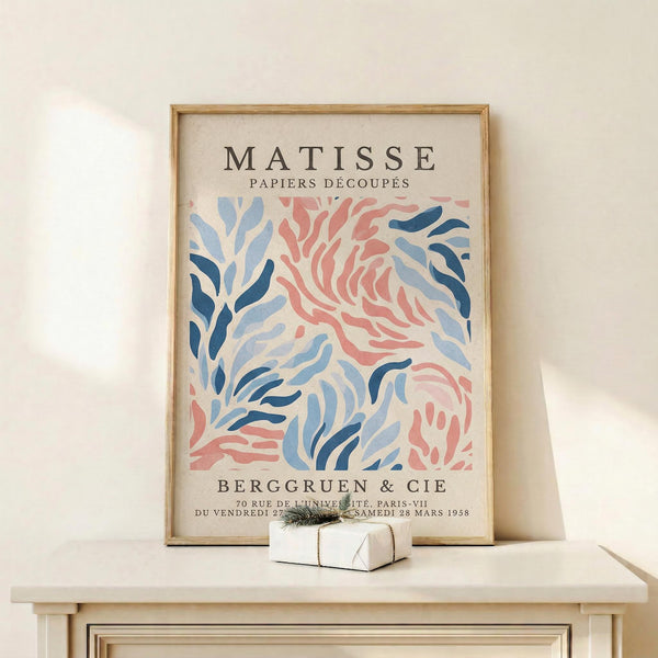 May include: Framed vintage-style exhibition poster featuring Matisse's cut-paper artwork in coral pink and blue organic flowing shapes against cream background, displayed on light wood console table beside small wrapped gift with greenery accent on white wall