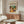 May include: A vintage-style Matisse exhibition poster featuring a colorful still life with wine bottle, fruits, and plants displayed above a mid-century modern walnut credenza. The room shows a white upholstered chair, glass oval coffee table, floor lamp, and neutral walls with natural lighting creating shadows.