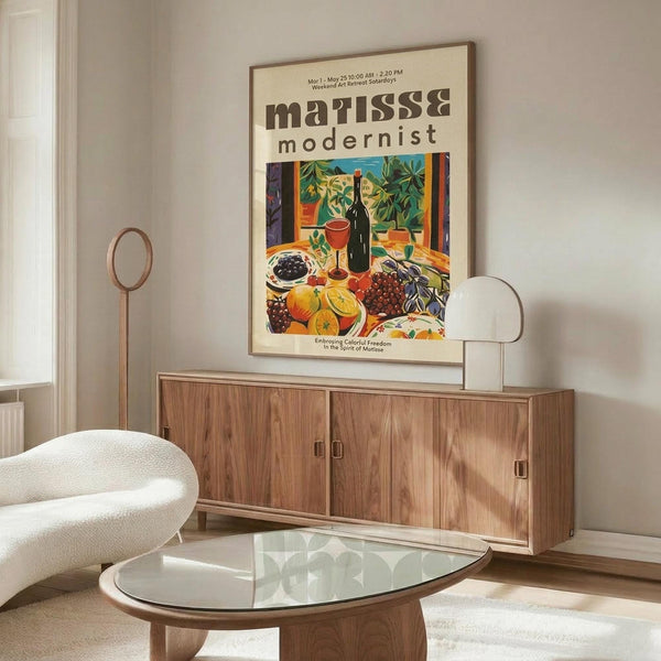 May include: A vintage-style Matisse exhibition poster featuring a colorful still life with wine bottle, fruits, and plants displayed above a mid-century modern walnut credenza. The room shows a white upholstered chair, glass oval coffee table, floor lamp, and neutral walls with natural lighting creating shadows.