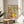 May include: Framed art poster displaying colorful botanical illustration with citrus fruits, leafy branches, bottles and bowls in vibrant yellows, greens, pinks and oranges. The artwork sits on a round wooden side table next to a beige upholstered chair with natural sunlight streaming across the neutral-toned room. A plant's leaves are visible in the corner, creating soft shadows on the white walls.