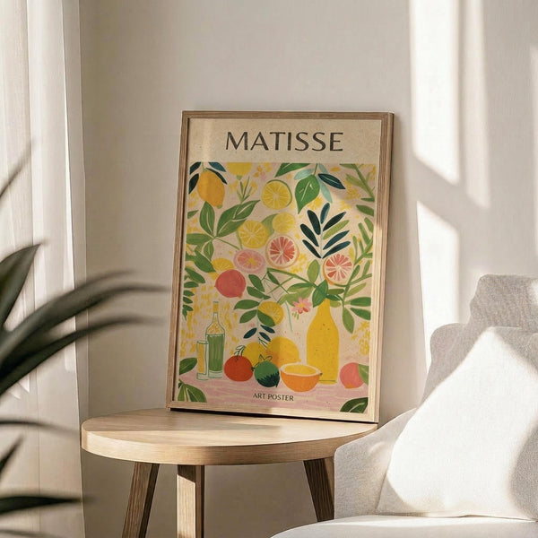 May include: Framed art poster displaying colorful botanical illustration with citrus fruits, leafy branches, bottles and bowls in vibrant yellows, greens, pinks and oranges. The artwork sits on a round wooden side table next to a beige upholstered chair with natural sunlight streaming across the neutral-toned room. A plant's leaves are visible in the corner, creating soft shadows on the white walls.
