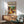 May include: Matisse-inspired modernist poster in wooden frame displaying colorful still life with wine bottle, fruits, and plants, positioned on mid-century wooden credenza beside white ceramic table lamp, next to sage green upholstered armchair with wooden arms in bright, sunlit room with white walls