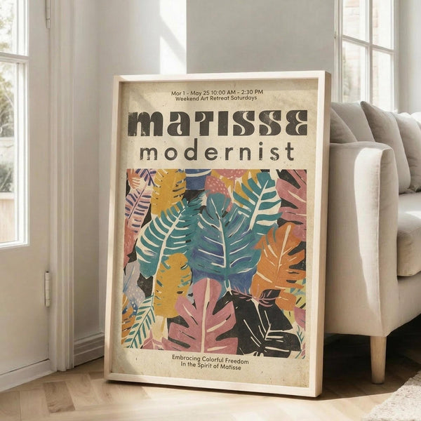 May include: Large framed art print leaning against white wall next to beige sofa in bright living room. Poster features colorful tropical leaves in pink, teal, orange, and yellow with bold black text reading "matisse modernist" and event details. Natural wood frame, vintage-style design on cream background.