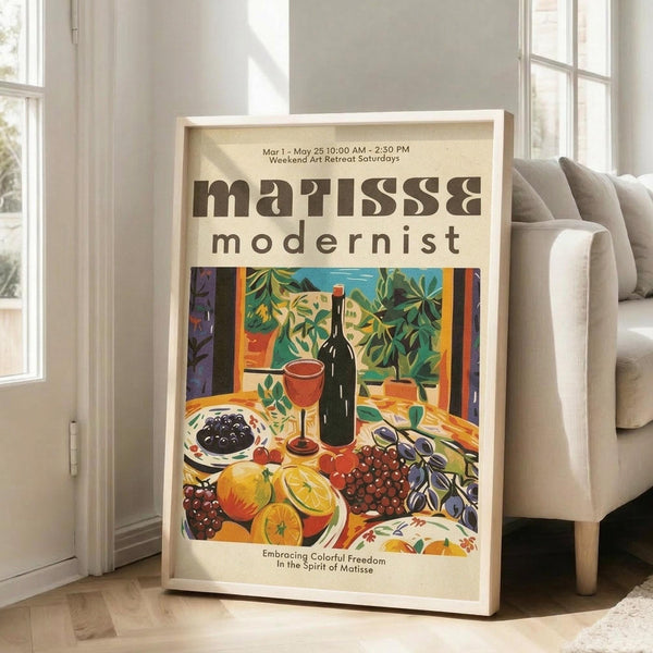 May include: Framed poster with cream background displaying colorful still life artwork in Matisse style. Features wine bottle, red wine glass, fruits including oranges and grapes, olives on plate, and lush green foliage. Bold oranges, reds, blues and greens create vibrant composition. Light wood frame leans against white wall next to beige sofa in bright room with large windows.