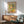 May include: Framed Matisse-style poster print hanging above wooden sideboard in modern living room. Artwork features colorful fruit still life with lemons, oranges, grapefruit slices, and green foliage in vibrant yellows, greens, and coral tones. Room includes sage green mid-century armchair with wooden arms, white ceramic table lamp, and natural sunlight casting shadows on white wall.