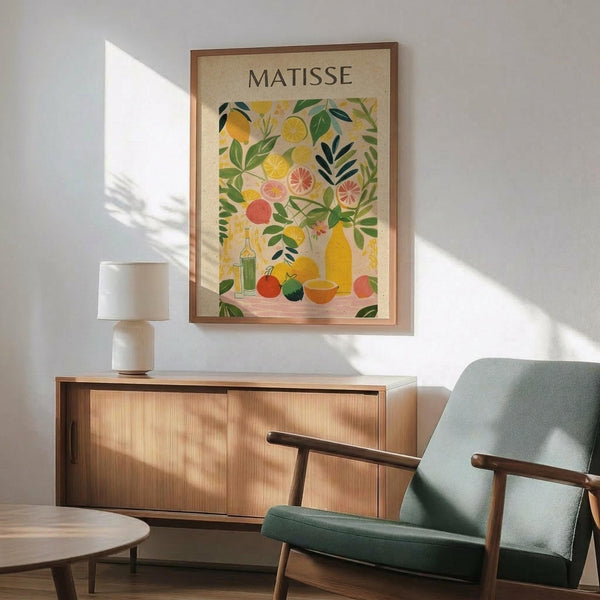 May include: Framed Matisse-style poster print hanging above wooden sideboard in modern living room. Artwork features colorful fruit still life with lemons, oranges, grapefruit slices, and green foliage in vibrant yellows, greens, and coral tones. Room includes sage green mid-century armchair with wooden arms, white ceramic table lamp, and natural sunlight casting shadows on white wall.