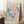 May include: Framed vintage-style exhibition poster featuring abstract coral pink and blue cut-out shapes in Matisse's papier découpé style, displayed leaning against white wall corner on fluffy cream rug next to wooden side table with white coffee cup and saucer, creating cozy modern living room vignette with natural window lighting.