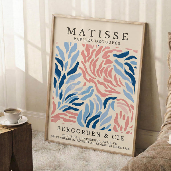 May include: Framed vintage-style exhibition poster featuring abstract coral pink and blue cut-out shapes in Matisse's papier découpé style, displayed leaning against white wall corner on fluffy cream rug next to wooden side table with white coffee cup and saucer, creating cozy modern living room vignette with natural window lighting.
