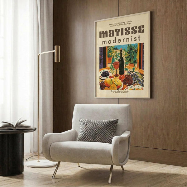May include: A framed vintage-style poster featuring a colorful still life with wine bottle, fruit, and foliage in Matisse-inspired style, displayed on wood-paneled wall above a beige upholstered armchair with geometric patterned pillow. Scene includes brass floor lamp and dark round side table with open book near window with white curtains.
