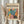 May include: Person holding framed modernist art poster featuring colorful botanical leaf cutouts in teal, pink, orange, and yellow against cream background. Light wood frame, dried pampas grass visible, neutral beige clothing and curtained backdrop create minimalist aesthetic.