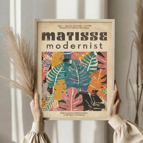 May include: Person holding framed modernist art poster featuring colorful botanical leaf cutouts in teal, pink, orange, and yellow against cream background. Light wood frame, dried pampas grass visible, neutral beige clothing and curtained backdrop create minimalist aesthetic.