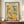 May include: Framed art poster in brown wooden frame displaying colorful still life with citrus fruits, bottles, and leafy plants in Matisse-inspired style. Warm yellows, greens, and coral tones on cream background. Sits on wooden console with white ceramic vase and stacked books in neutral-toned room with white paneled walls.