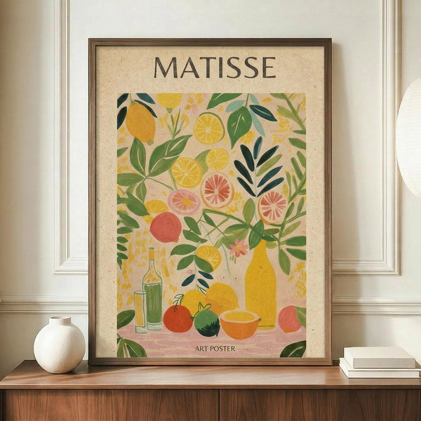 May include: Framed art poster in brown wooden frame displaying colorful still life with citrus fruits, bottles, and leafy plants in Matisse-inspired style. Warm yellows, greens, and coral tones on cream background. Sits on wooden console with white ceramic vase and stacked books in neutral-toned room with white paneled walls.