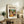 May include: A framed poster featuring a colorful still life scene with fruits, wine bottle, and foliage in vibrant greens, oranges, and reds. The artwork displays "matisse modernist" text at the top with event details. The wooden frame sits on a rustic console table alongside books, a ceramic vase, and a fern plant in a cozy living room setting with neutral tones and soft lighting.