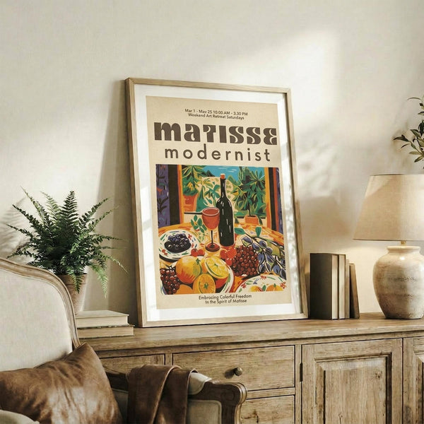 May include: A framed poster featuring a colorful still life scene with fruits, wine bottle, and foliage in vibrant greens, oranges, and reds. The artwork displays "matisse modernist" text at the top with event details. The wooden frame sits on a rustic console table alongside books, a ceramic vase, and a fern plant in a cozy living room setting with neutral tones and soft lighting.