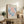 May include: Framed vintage-style exhibition poster featuring Matisse's colorful cut-out designs in coral pink and blue tones, displayed on rustic wooden sideboard alongside fern plant, ceramic vase, stacked books, and table lamp in warm neutral living room setting