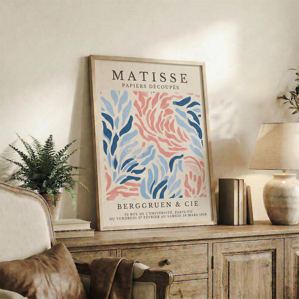May include: Framed vintage-style exhibition poster featuring Matisse's colorful cut-out designs in coral pink and blue tones, displayed on rustic wooden sideboard alongside fern plant, ceramic vase, stacked books, and table lamp in warm neutral living room setting
