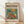 May include: Framed poster in natural wood frame leaning against white wall on light wooden floor. Cream-colored poster features "matisse modernist" text and colorful illustration of orange goldfish swimming in glass fishbowl with bright orange and yellow flowers, green leaves on teal background. Soft natural lighting creates diagonal shadows across white fabric backdrop.