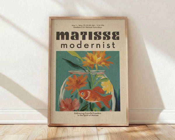 May include: Framed poster in natural wood frame leaning against white wall on light wooden floor. Cream-colored poster features "matisse modernist" text and colorful illustration of orange goldfish swimming in glass fishbowl with bright orange and yellow flowers, green leaves on teal background. Soft natural lighting creates diagonal shadows across white fabric backdrop.