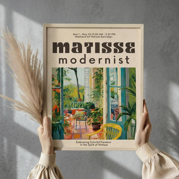 May include: Hands holding a framed art poster featuring a vibrant interior scene with yellow chair, potted plants, and view through French doors to a garden balcony. The poster has "matisse modernist" text and cream background. Dried pampas grass visible on left side against textured gray wall with natural lighting.
