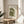 May include: Framed green and beige art print featuring an abstract seated figure in Matisse's cut-out style, displayed on a round wooden side table next to a beige upholstered chair. Soft natural lighting creates shadows on white walls, with plant leaves visible in the corner of the minimalist room setting.