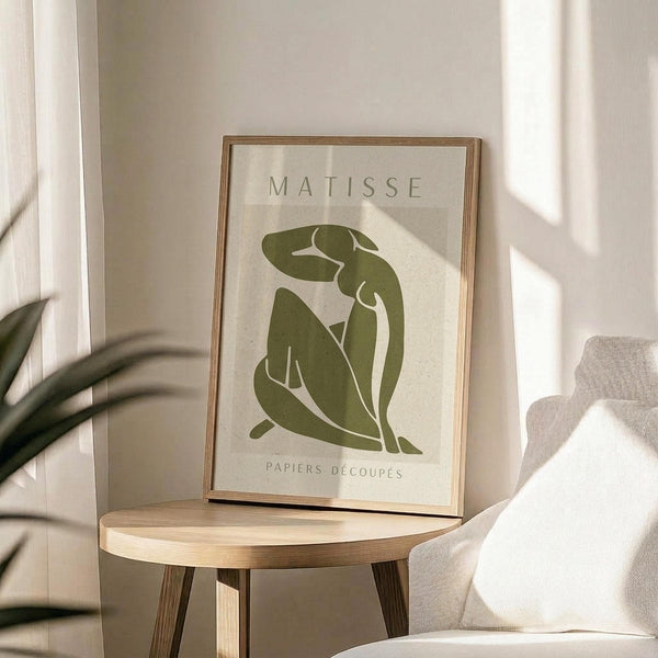 May include: Framed green and beige art print featuring an abstract seated figure in Matisse's cut-out style, displayed on a round wooden side table next to a beige upholstered chair. Soft natural lighting creates shadows on white walls, with plant leaves visible in the corner of the minimalist room setting.