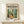 May include: Framed art print featuring colorful modernist interior scene with lush green plants, yellow chair, and blue window frames in Matisse-inspired style. Print displayed in light wood frame on white wooden surface beside small wrapped gift with twine and greenery sprig against cream wall.