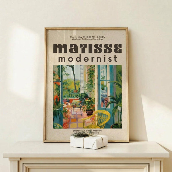 May include: Framed art print featuring colorful modernist interior scene with lush green plants, yellow chair, and blue window frames in Matisse-inspired style. Print displayed in light wood frame on white wooden surface beside small wrapped gift with twine and greenery sprig against cream wall.