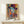 May include: Colorful portrait poster in wooden frame showing woman wearing ornate hat, rendered in vibrant blues, oranges, purples and yellows with decorative patterns. Artwork displays "MATISSE" text at bottom. Frame sits on light wood surface next to small wrapped white gift box with green sprig against cream wall.