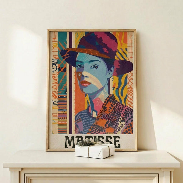 May include: Colorful portrait poster in wooden frame showing woman wearing ornate hat, rendered in vibrant blues, oranges, purples and yellows with decorative patterns. Artwork displays "MATISSE" text at bottom. Frame sits on light wood surface next to small wrapped white gift box with green sprig against cream wall.