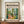 May include: Framed vintage-style art poster featuring colorful interior scene with plants, yellow chair, and windows, displayed on wooden console with white ceramic vase and books against cream paneled wall