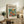 May include: Framed poster with cream matting displaying colorful modernist artwork of orange goldfish swimming among vibrant flowers in glass vase against teal background. Print sits on rustic wooden sideboard beside fern plant, ceramic vases, and books in warmly lit living room with beige sofa and neutral decor.
