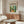 May include: Modernist art poster featuring colorful interior scene with plants, furniture and windows, displayed above wooden credenza in contemporary living room setting with white curved chair, glass coffee table, and modern floor lamp