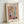 May include: Framed art poster in light wood frame leaning against white wall. Print features Matisse-style cut-out artwork depicting colorful window scene with geometric panels, plant motifs in green, blue, orange and pink tones, dark blue vase, and potted plant. Text reads "MATISSE" at top and "ART POSTER" at bottom on cream background.