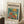 May include: Framed poster with Matisse-inspired artwork featuring an orange goldfish swimming among colorful flowers in a glass bowl against a teal background. The vintage-style print shows "modernist" text and is displayed leaning against white wainscoting on a textured cream rug, with a coffee cup on a wooden block nearby and cozy beige throw blanket visible.