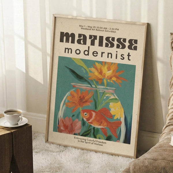 May include: Framed poster with Matisse-inspired artwork featuring an orange goldfish swimming among colorful flowers in a glass bowl against a teal background. The vintage-style print shows "modernist" text and is displayed leaning against white wainscoting on a textured cream rug, with a coffee cup on a wooden block nearby and cozy beige throw blanket visible.