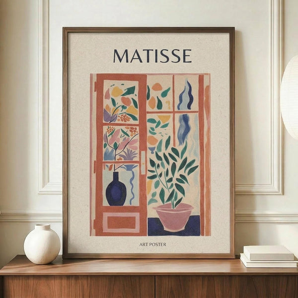 May include: Framed Matisse-style art poster displaying colorful cut-out inspired window scene with plants, blue vase, and botanical elements in warm terracotta tones. Wooden frame sits on walnut credenza beside white ceramic vase and stacked books against cream wainscoted wall.