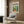 May include: Cream-colored framed Matisse modernist art print hanging on wood-paneled wall above beige upholstered armchair with geometric patterned pillow. Print features vibrant interior scene with green plants, colorful furniture, and windows. Gold floor lamp and dark side table with open book complete the modern living room setting.