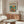 May include: Matisse-inspired modernist poster displayed above wooden sideboard in bright living room. Artwork features colorful goldfish in glass bowl with orange and yellow flowers against teal background. White upholstered chair and glass coffee table visible. Natural wood furniture with mid-century design. Soft natural lighting creates warm, contemporary interior styling.