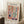 May include: Framed Matisse-style art poster leaning against white wall corner on textured rug. Poster features colorful cutout-style window scene with orange frame, blue vase, green plant in pink pot, and abstract botanical elements in yellow, coral, and teal. Light wood frame, coffee cup on wooden block nearby, beige throw blanket visible.