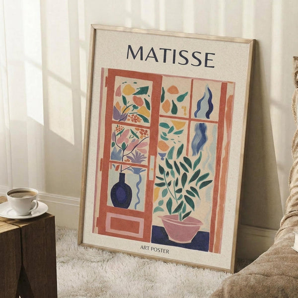 May include: Framed Matisse-style art poster leaning against white wall corner on textured rug. Poster features colorful cutout-style window scene with orange frame, blue vase, green plant in pink pot, and abstract botanical elements in yellow, coral, and teal. Light wood frame, coffee cup on wooden block nearby, beige throw blanket visible.