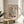 May include: Framed Matisse art poster featuring colorful cut-out style illustrations of plants, vases, and decorative elements in coral, blue, green, and pink tones. The print sits on a light wood round side table next to a beige upholstered chair, with natural sunlight streaming across neutral walls and a plant visible in the corner.