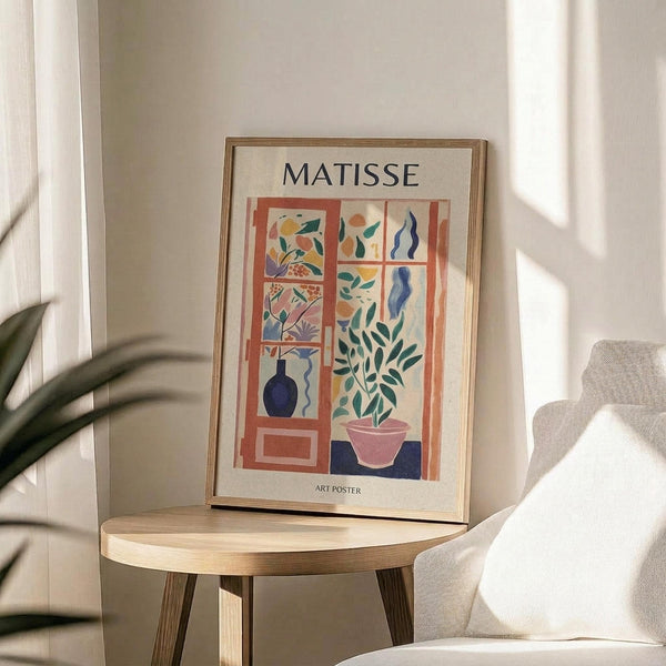 May include: Framed Matisse art poster featuring colorful cut-out style illustrations of plants, vases, and decorative elements in coral, blue, green, and pink tones. The print sits on a light wood round side table next to a beige upholstered chair, with natural sunlight streaming across neutral walls and a plant visible in the corner.
