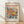 May include: Framed poster print leaning against white wall on wooden floor. Cream-colored poster features vibrant floral artwork with coral, yellow, and blue flowers and green foliage in Matisse's cut-out style. Red "MATISSE" text at top, museum exhibition details below. Natural wood frame, soft lighting creates diagonal shadows on textured white wall.