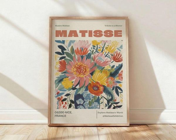 May include: Framed poster print leaning against white wall on wooden floor. Cream-colored poster features vibrant floral artwork with coral, yellow, and blue flowers and green foliage in Matisse's cut-out style. Red "MATISSE" text at top, museum exhibition details below. Natural wood frame, soft lighting creates diagonal shadows on textured white wall.