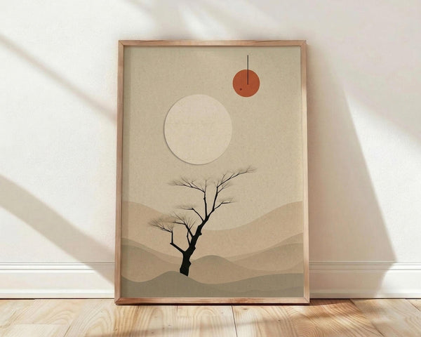 May include: Framed minimalist desert landscape artwork leaning against white wall on light wood floor. Art features layered beige and tan hills, bare black tree silhouette, large cream circle and smaller rust orange circle representing celestial bodies with thin black line. Natural wood frame with warm shadows cast across scene.
