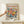 May include: Person holding a framed colorful floral poster in light wood frame, featuring vibrant pink, yellow, orange and blue flowers with green leaves in Matisse's cut-out style. Poster displays "MATISSE" in bold red letters at top, museum information, and social media handle at bottom. Small wrapped gift box with twine and greenery sits on light wooden surface below the frame against cream wall.