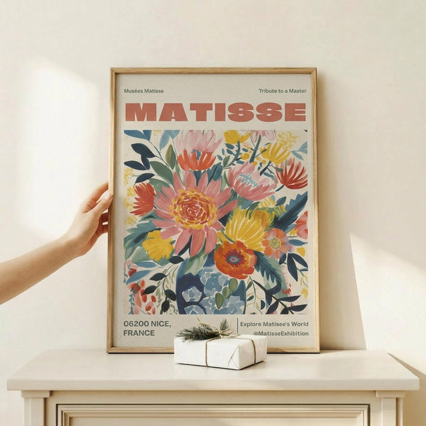 May include: Person holding a framed colorful floral poster in light wood frame, featuring vibrant pink, yellow, orange and blue flowers with green leaves in Matisse's cut-out style. Poster displays "MATISSE" in bold red letters at top, museum information, and social media handle at bottom. Small wrapped gift box with twine and greenery sits on light wooden surface below the frame against cream wall.