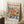 May include: A framed museum exhibition poster featuring vibrant watercolor-style flowers in coral, yellow, and blue tones against a cream background. The wooden frame leans against white paneled walls on a textured white rug, with a coffee cup on a wooden stool nearby and cozy fabric visible at the edge.